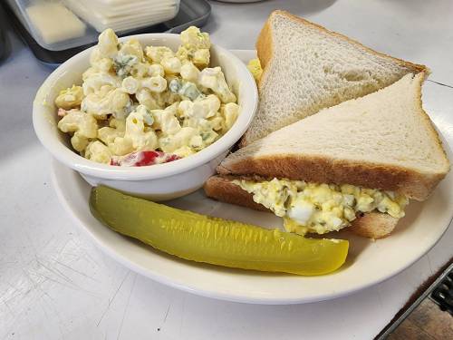 A plate with a sandwich filled with egg salad, a side of macaroni salad, and a pickle slice.
