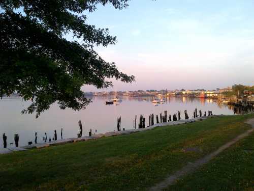 A serene waterfront scene with boats, wooden pilings, and a grassy shore under a pastel sky.