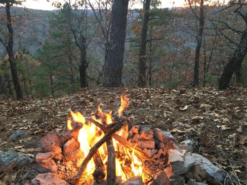 A campfire burns among rocks and leaves, surrounded by trees and a scenic view of a forested landscape at sunset.