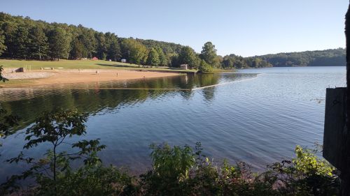 A serene lake surrounded by trees, with a sandy beach and clear blue sky reflecting on the water's surface.