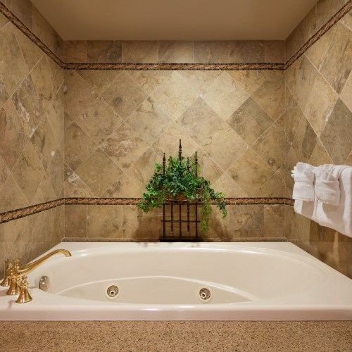 A luxurious bathroom featuring a white jacuzzi tub, stone walls, and decorative greenery. Soft towels are neatly arranged.
