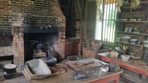 A rustic kitchen with a brick fireplace, wooden table, and various cooking utensils and pottery on display.