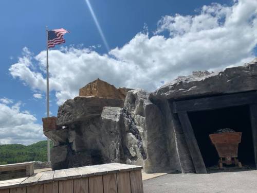 A rocky landscape with an American flag, a mine entrance, and a coal cart under a partly cloudy sky.