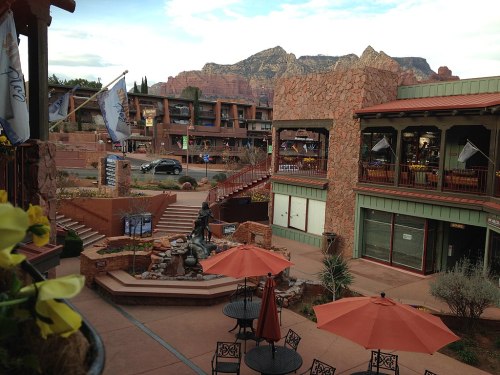 A scenic view of a shopping area with outdoor seating, a fountain, and mountains in the background.