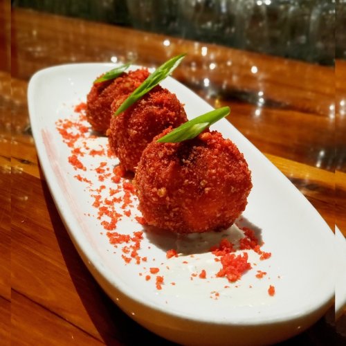 Three crispy, red-coated balls garnished with green onions, served on a white plate with a sprinkle of red crumbs.