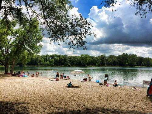 A sandy beach by a lake with people swimming and relaxing under trees and clouds.