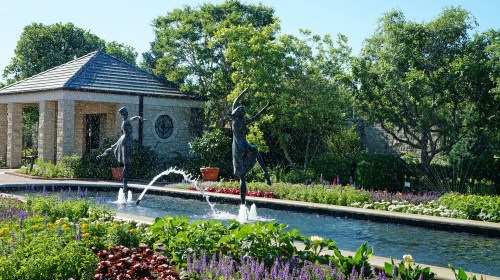 A serene garden scene featuring two dancing sculptures by a fountain, surrounded by colorful flowers and greenery.