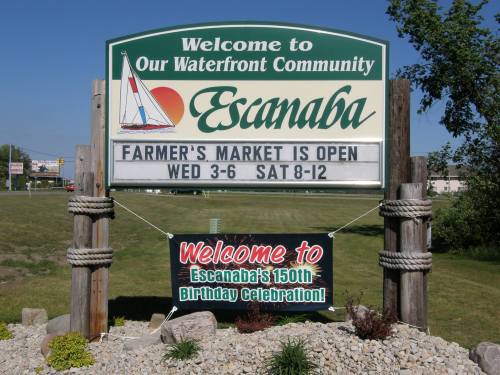 Sign welcoming visitors to Escanaba, featuring details about the farmer's market and a celebration for its 150th birthday.