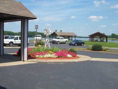 A scenic view of a parking area with flower beds, a windmill, and a lake in the background under a clear blue sky.