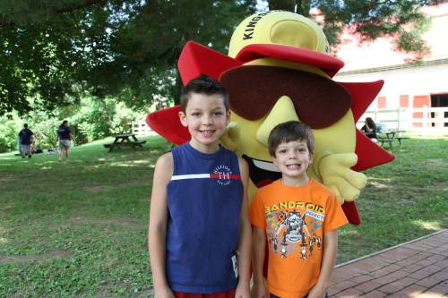 Two young boys smile for a photo with a large, cheerful sun mascot in a park setting.