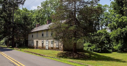 Historic stone house surrounded by trees, located near a road on a sunny day.