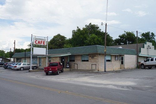 A small cafe building with a green roof and sign, surrounded by parked cars and trees under a cloudy sky.