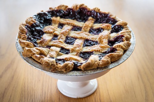 A cherry pie with a lattice crust, sitting on a white pedestal against a wooden table background.