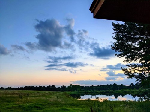 A serene landscape featuring a lake, grassy field, and a colorful sky with clouds at sunset.