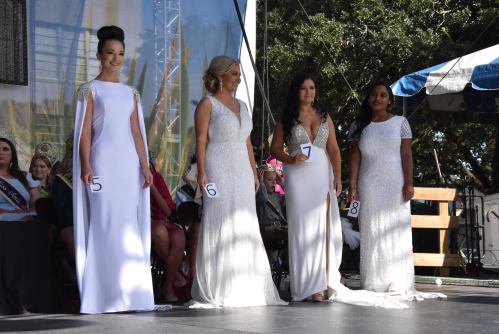 Four women in elegant white gowns stand on stage, each wearing a number, during a pageant event.