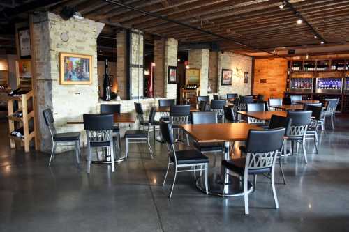 A modern restaurant interior featuring wooden tables, black chairs, and a wine display in the background.