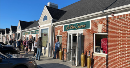 A brick storefront with a sign reading "Le Bon Sucre," featuring people waiting outside on a sunny day.