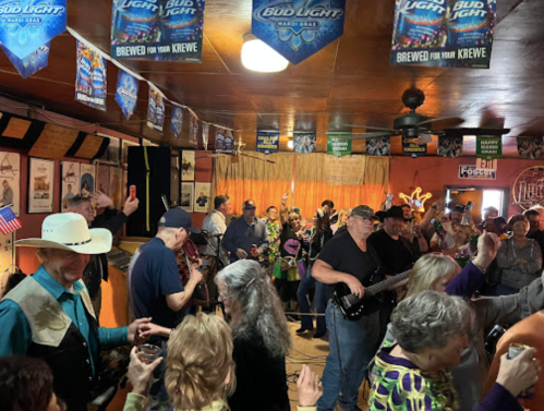 A lively crowd dances and enjoys music in a vibrant bar, decorated with beer banners and colorful lights.