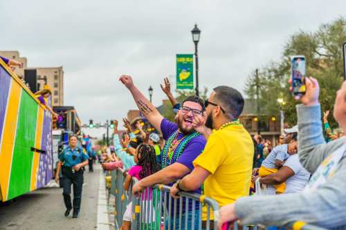 A lively parade scene with people celebrating, wearing beads, and cheering along the street.