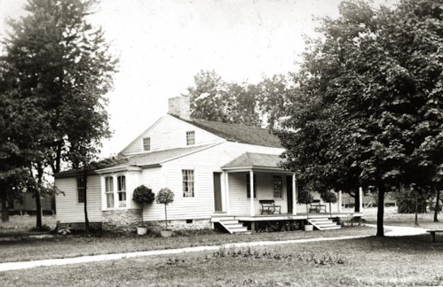 A vintage black-and-white photo of a modest house with a porch, surrounded by trees and a grassy yard.