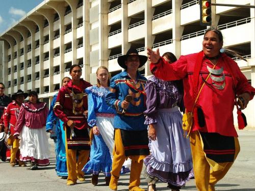 A group of people in traditional attire joyfully march together in a parade, with a parking garage in the background.