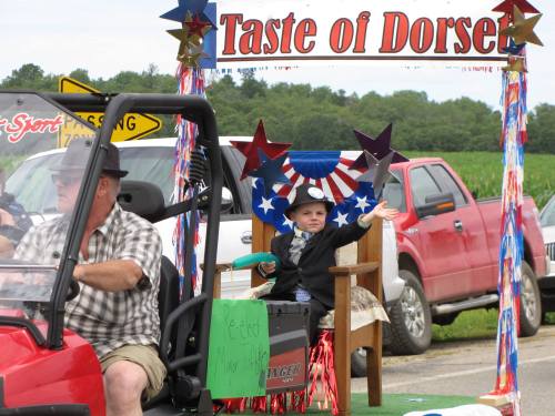 A young boy in a suit waves from a decorated vehicle in a parade, with a sign reading "Taste of Dorsey" above him.