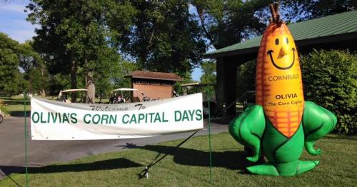 A banner reading "Welcome to Olivia's Corn Capital Days" next to a large inflatable corn character named Cornelius.