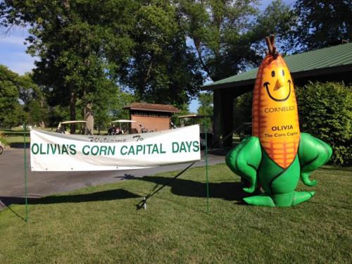 A banner reading "Welcome to Olivia's Corn Capital Days" with a large inflatable corn character nearby.