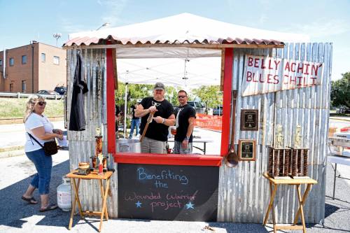Two men stand behind a booth decorated with trophies, promoting a chili event benefiting the Wounded Warrior Project.