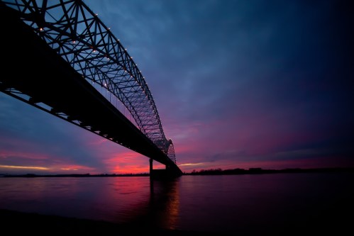 A silhouette of a bridge against a vibrant sunset, reflecting colors on the water below.