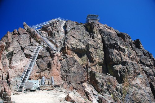 A steep rocky mountain with a lookout tower at the top and metal stairs leading up the side against a clear blue sky.