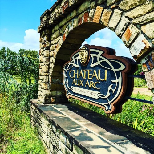 Sign for Chateau Aux Arc Vineyards & Winery, framed by a stone arch, surrounded by greenery and a blue sky.