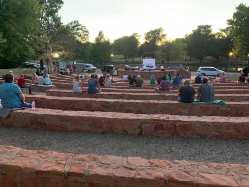 People sitting in an outdoor amphitheater watching a movie on a screen as the sun sets.