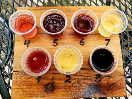 A wooden flight board with seven cups of different colored beverages arranged in a row.