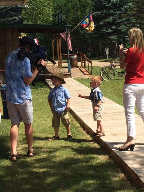 Two young boys in summer outfits stand on a path while a cameraman films them; a woman takes photos nearby.