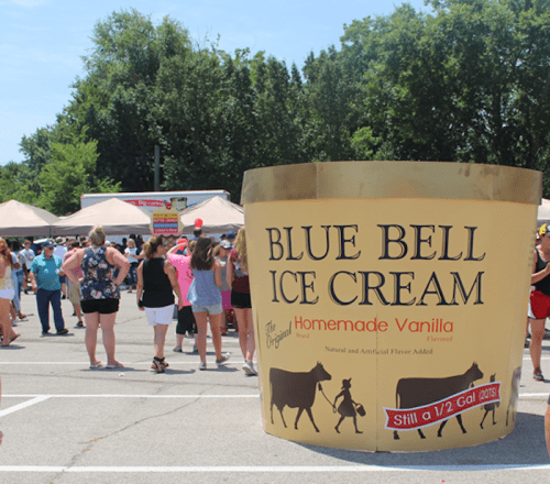 A large Blue Bell Ice Cream container stands in a crowded outdoor event with people enjoying the festivities.