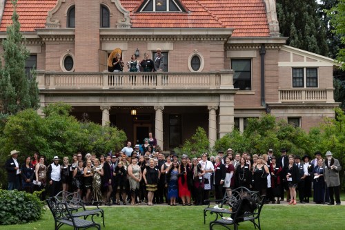 A large group of people in formal attire poses in front of a historic mansion, surrounded by greenery.