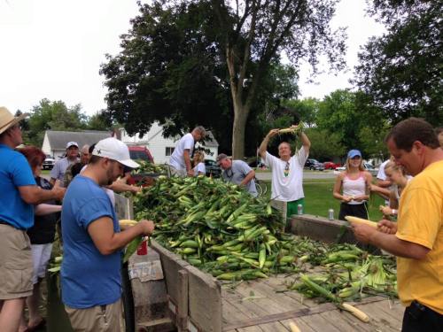 A group of people gather around a truck filled with corn, preparing it for an event in a sunny outdoor setting.