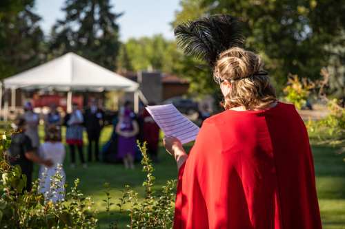 A person in a red cape reads from a paper, with guests in formal attire gathered outdoors under a tent.