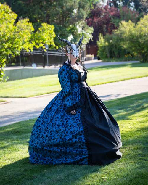 A person in a blue and black gown with horns poses in a lush garden setting.