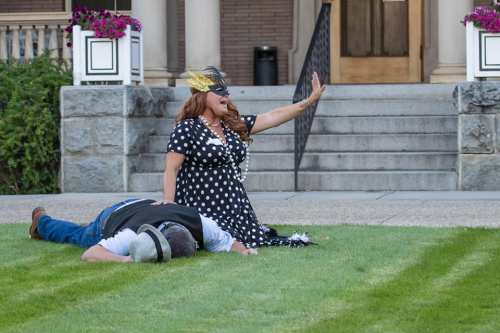 A woman in a polka dot dress and mask gestures dramatically while a man lies face down on the grass.