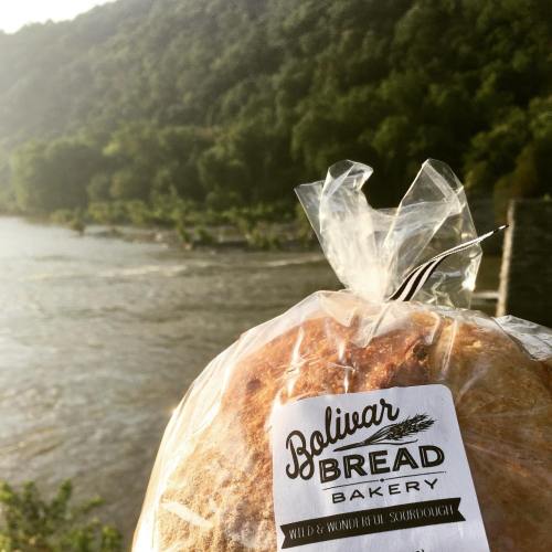 A loaf of sourdough bread in a clear bag, with a scenic river and mountains in the background.