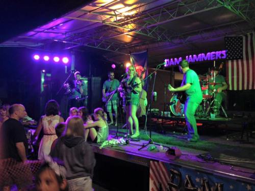 A lively band performs on stage with colorful lights, while an audience enjoys the music in front of them.