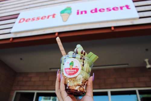 A hand holds a dessert cup with ice cream and toppings in front of a sign that reads "Dessert in Desert."