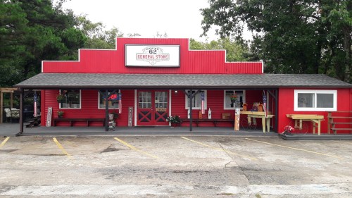 A red general store with a sign reading "Route 62 General Store," featuring a porch and decorative plants.