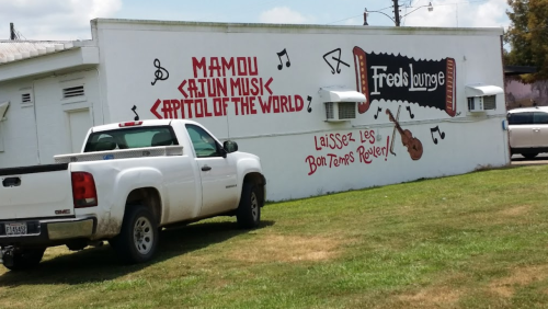 A white wall with a mural reading "Mamou Cajun Music Capital of the World" and "Fred's Lounge," with musical notes and a guitar.