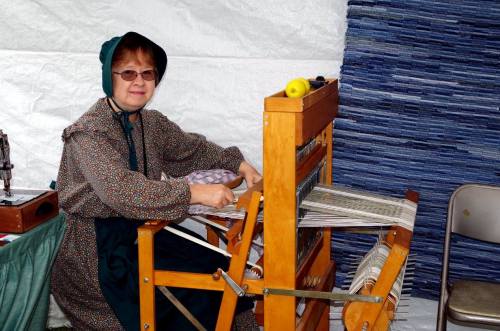 A woman in a bonnet weaves at a loom, surrounded by fabric and a white backdrop.