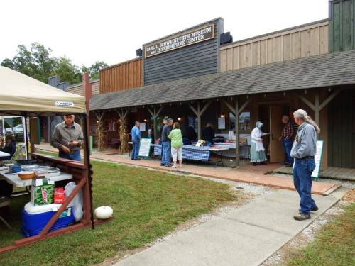 A group of people gather outside a museum with a rustic facade, featuring tables and tents for an event.