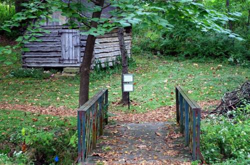 A rustic wooden bridge leads to a cabin in a wooded area, surrounded by trees and fallen leaves.