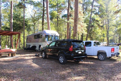 A large bus parked among trees, with a black SUV and a white pickup truck nearby in a wooded area.